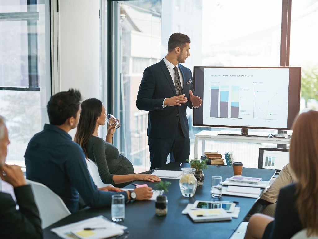 User-Inserted Business Meeting With A Presenter Explaining Sales Data On A Screen, While Colleagues Listen And Take Notes In A Modern Office Setting.
